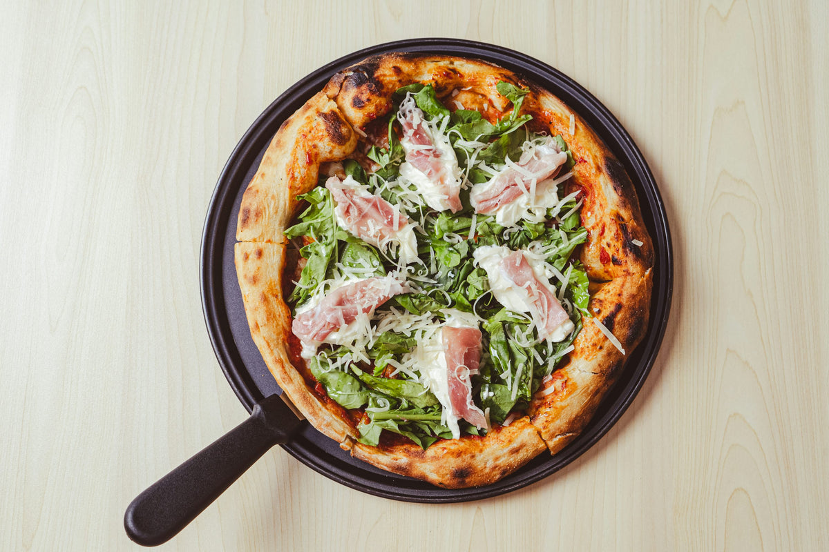 Servieridee: Runde Pizza mit Rohschinken, Rucola und hellen Käsespänen auf einem dunklen Pizzabrett. Foto: Aldward Castillo / Unsplash.