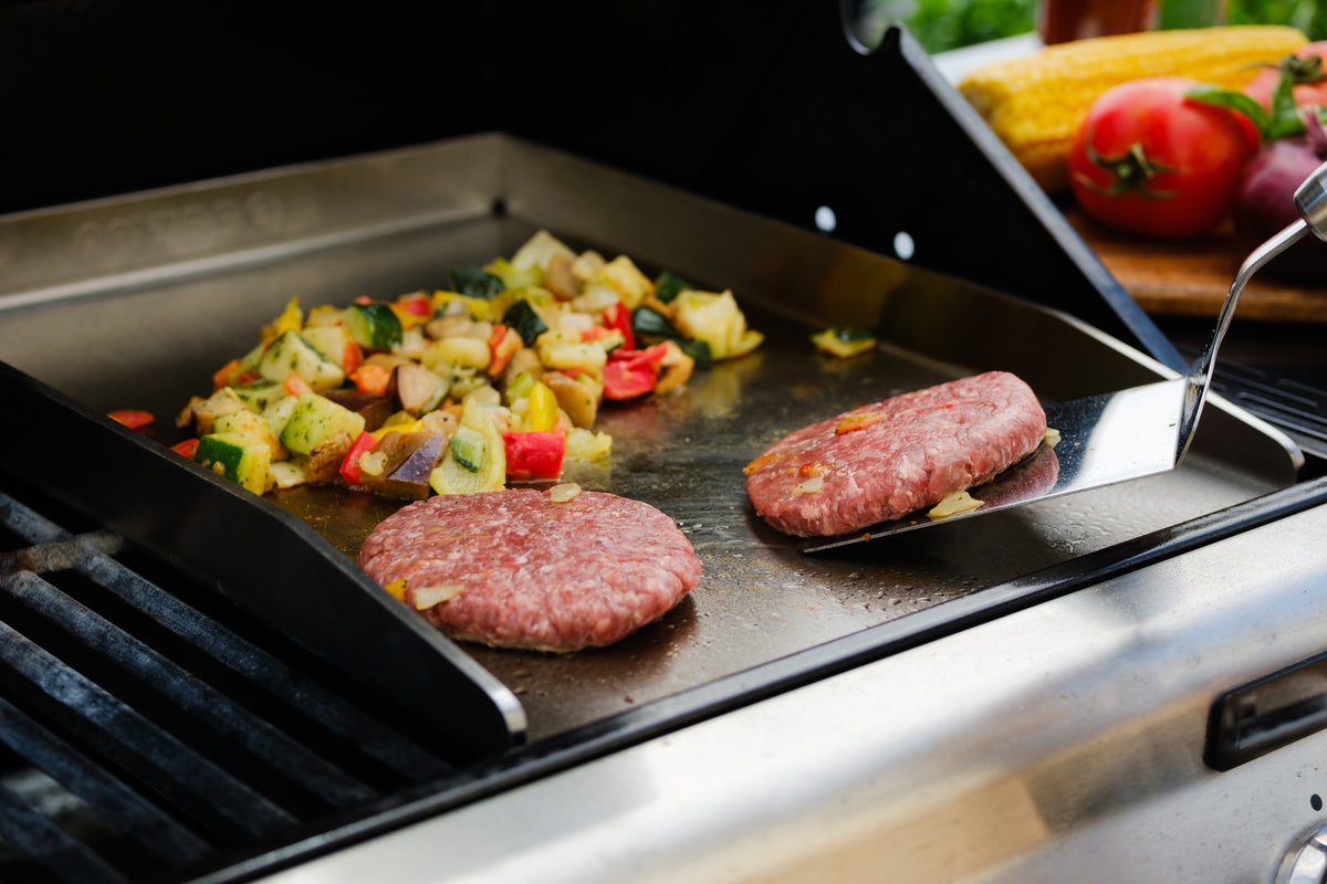 Burger und Gemüse auf einer Edelstahlplancha neben dem Grillrost
